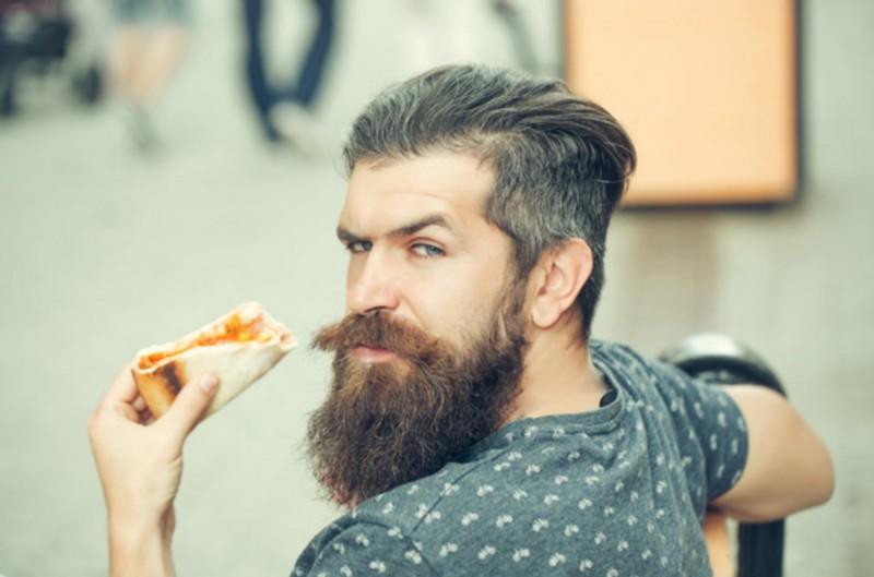 Hipster man with a beard staring you down as he eats pizza.