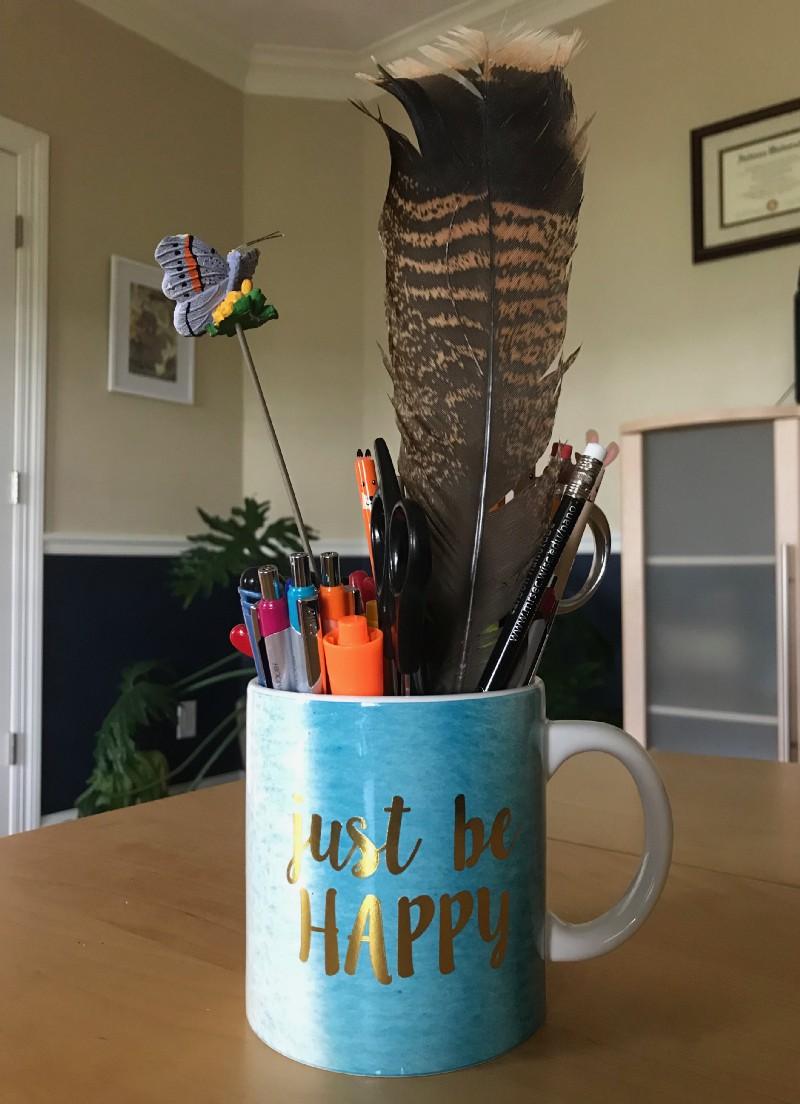 Just be happy mug filled with pens, pencils, and my turkey feather.
