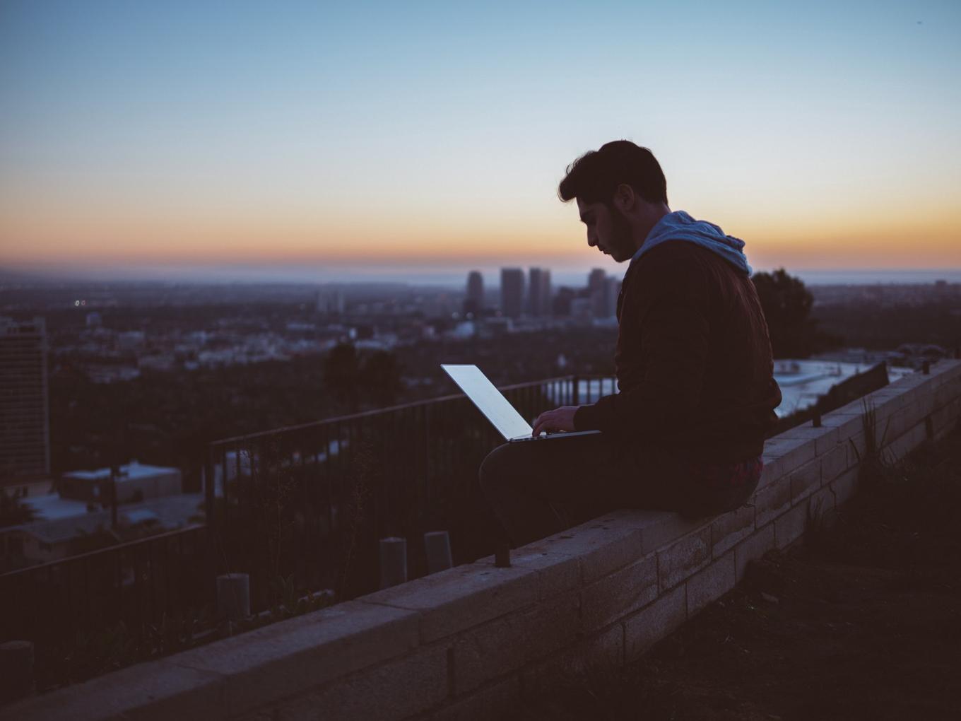 Man sitting on a building ledge at dusk trying to use his computer.